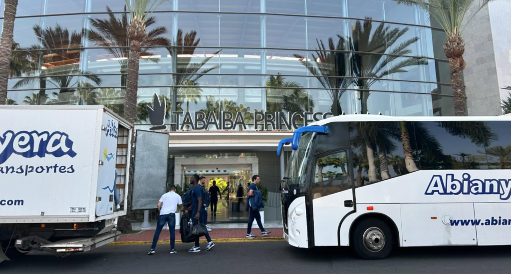 Autobús y camión de transporte frente a la entrada del hotel Tabaiba Princess, con varias personas cargando equipaje